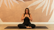 A woman sits on a yoga mat. She is wearing dark yoga leggings and black athletic tank top. She has shoulder-length brown hair and her eyes are closed peacefully. She has a slight smile on her face and has placed her hands over her chest. 
