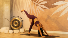 A woman in athletic wear practices yoga on a mat, holding a downward dog split pose with one leg raised high. She is in a calm studio with warm-toned walls, leaf-pattern decor, and a set of white singing bowls and a gong placed nearby.