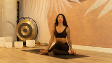 A woman sits cross-legged on a yoga mat in a meditation pose with eyes closed, wearing black athletic clothing. She is in a warm, softly lit studio with a leaf-pattern wall mural, and a gong and white singing bowls arranged nearby.