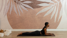 A woman in black athletic wear lies on her stomach on a yoga mat in a cobra pose, propped on her forearms and looking to the side. The studio has a warm-toned wall with a large leaf mural, and white singing bowls are arranged nearby.