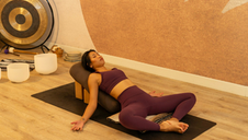 A woman in purple workout clothes reclines on a yoga mat in a butterfly stretch position, leaning back on a cylindrical bolster with her arms relaxed at her sides in a warm-toned studio.