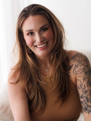Rachel Vanoven, photographer and educator, smiling with long hair, hoop earrings, and visible tattoos, seated against a light background.