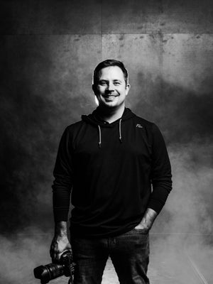 G Paschal, Sportraits photographer, smiling in a black hoodie against a dark textured studio backdrop.