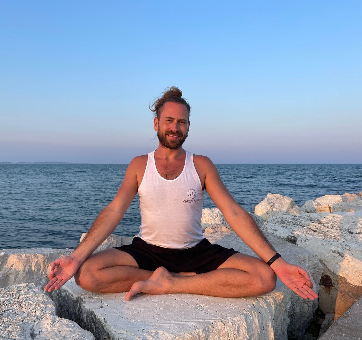 Andrew smiling, seated cross-legged on white rocks by the sea at sunset, arms open