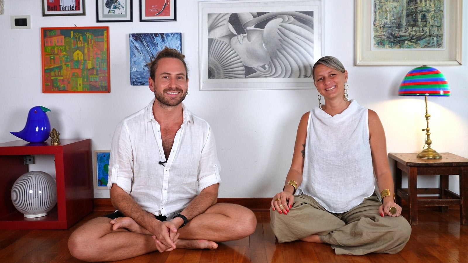 Andrew and Clelia smiling, sitting cross-legged on a wooden floor in front of a wall of paintings
