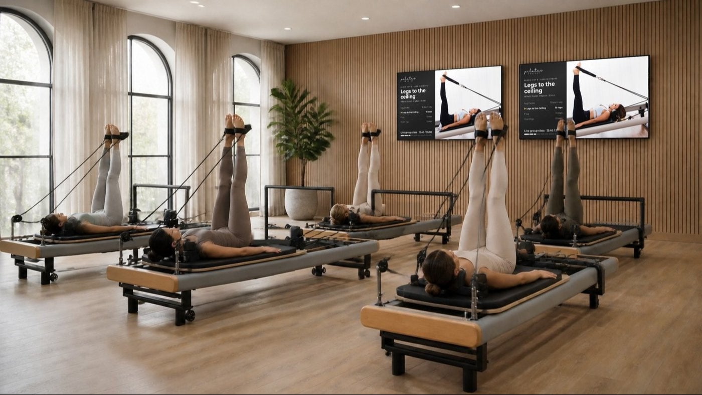 Members on reformers doing Legs to the Ceiling in straps while two wall-mounted screens show the same exercise